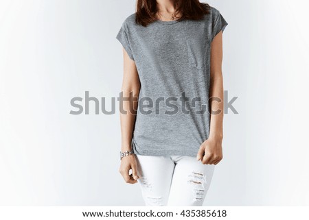 Fashionable young Caucasian woman posing indoor against white wall background. Cropped shot of hipster teenage girl wearing blank T-shirt and stylish ripped jeans. Lifestyle and people concept.