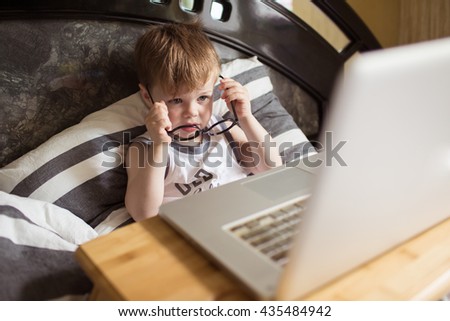 Toddler boy in glasses laying on the bed and loooking to pc at home, indoors. Child with computer