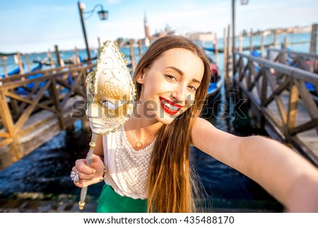 Young female traveler making selfie photo with carnaval mask standing near San Marco square with gondolas on the background in Venice.