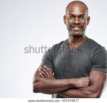 Single handsome fit grinning young Black adult with shaved head and folded muscular arms over gray background with copy space