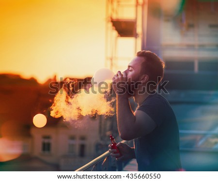 Man with beard smoking vaporizer outdoor and making soap bubbles with steam inside, against the backdrop of the sunset sky
