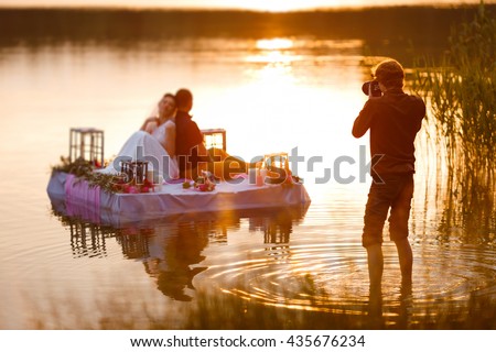 Wedding photographer in action, taking a picture of the bride and groom sitting on the raft. Summer, sunset Royalty-Free Stock Photo #435676234