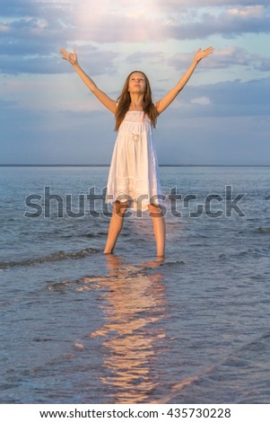 Girl in a white sundress standing in the water at the beach with their hands raised in the last rays of the sun and says goodbye to the setting sun