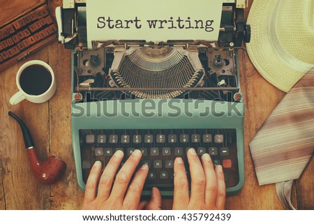 top view photo of man typing on vintage typewriter with text: START WRITING next to cup of coffee and man accessories, on wooden table. retro filtered image