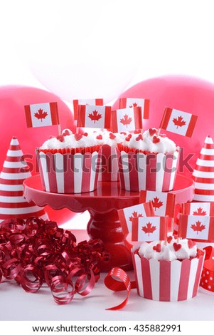 Happy Canada Day Party Cupcakes on a red cake stand with maple leaf flags on a white wood table with party balloons, hats and decorations.