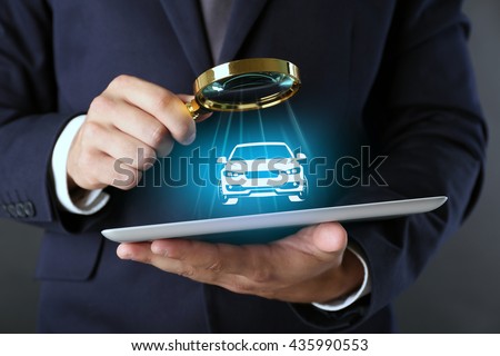Businessman holding magnifying glass and digital tablet with virtual car on dark grey background