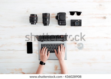 Hands of young man using laptop on wooden table with blank screen mobile phone, glass of water, sunglasses, lenses and photo camera