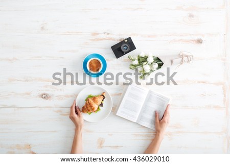 Hands of young woman eating croissant with coffee and reading book on wooden table with flower bouquet and photo camera