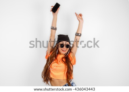 beautiful woman in sunglasses wearing in black hat and orange T-shirt listening music and dancing near white wall, holding a cell phone in hand, hands raised up