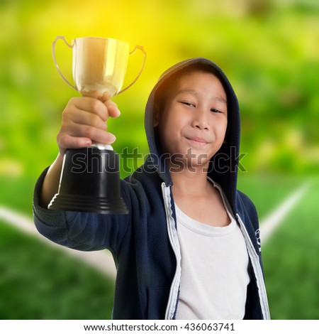 Happy boy in blue hood jacket holding trophy with soccer field background