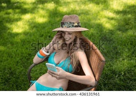 Young woman wearing bikini and sunhat reading smartphone texts in garden