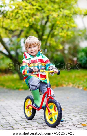 Cute blond kid boy driving on bicycle on rainy summer or autumn day outdoors. Preschool child  in colorful rain jacket and boots having fun in park.