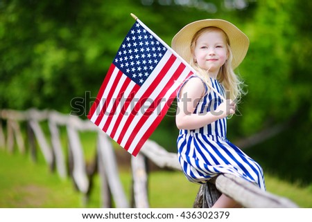 Adorable little girl wearing hat holding american flag outdoors on beautiful summer day. Independence Day concept.