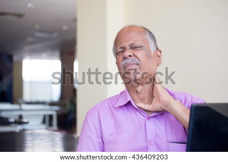 Closeup portrait, elderly gentleman in pink shirt with cramping neck pain after using black laptop all day, trying to soothe with hand, isolated indoors background