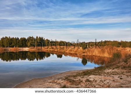 Evening on the pond, beach and forest, blue sky with clouds and reflection in water.