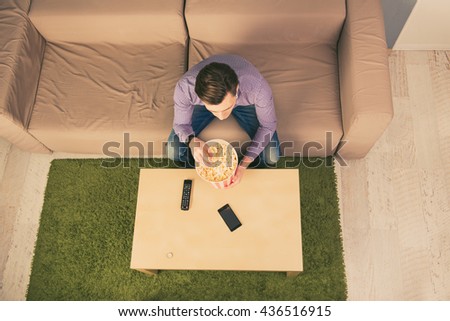 Top view of young man resting at home while watching film and eating popcorn