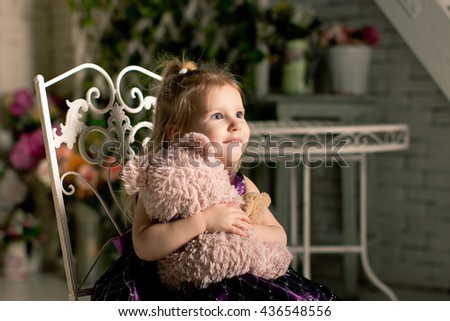 The little blonde girl with Teddy bear  looking forward. The face is beautifully lit. The girl is looking ahead and is excited about it. She sits on a beautiful vintage white chair