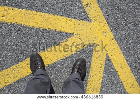 Male feet in leather shoes stand on on asphalt pavement with yellow road marking lines, first person view