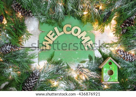 Welcome sign with green Christmas tree garland border, lights, pine cones and birdhouse on antique rustic snowy wood background