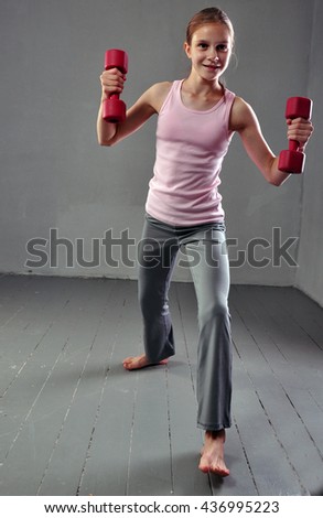 Teenage sportive girl is doing exercises with dumbbells to develop muscles on grey background. Sport healthy lifestyle concept.  Full length portrait of teenager child exercising with weights.