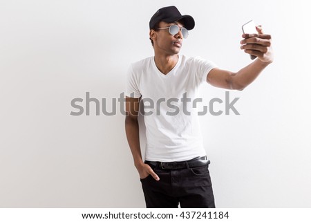 Casual black guy in shades and cap taking selfie with smart phone on light background with copy space