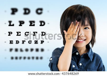 Close up portrait of cute little asian boy doing eye test.Kid closing one eye with hand against alphabetical out of focus test chart in background.