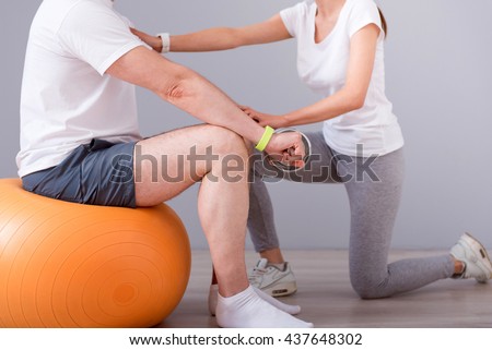 Rehabilitation together. Cropped image of male patient sitting on a gym ball with dumbbells during physical therapy praxis and female physiotherapist exercising with him Royalty-Free Stock Photo #437648302