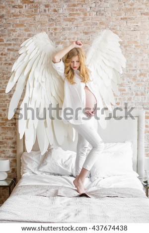Young beautiful red-haired pregnant girl in a white shirt and jeans with wings behind the back, the angel, a home style that embraces the belly, look out the window, tenderness, purity, health