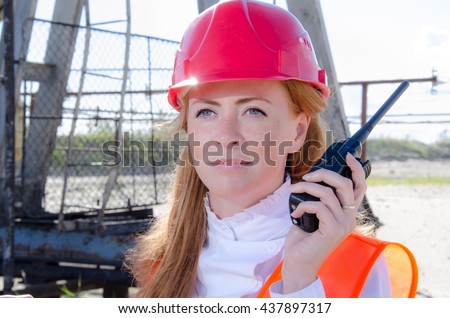 Beautiful woman engineer in the oil field talking on the radio wearing red helmet and work clothes. Pump jack background. Oil and gas concept. 