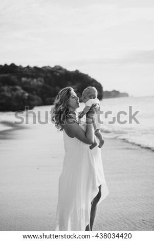 young smiling mother in flying white dress with her baby on the beach . Mother holds newborn baby in her arms . Happy woman with a child smiling and kissing her baby. close up portrait out door