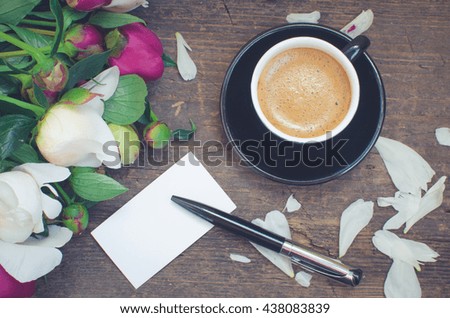 A cup of coffee and flowers Peonies on a table with an empty card for text and pen. Breakfast on Mothers day, Valentines Day or Womens day. Writing greetings card. Copy space. Top view. Toned image.