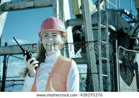 Beautiful woman engineer in the oil field talking on the radio wearing red helmet and work clothes. Pump jack background. Oil and gas concept. Toned.