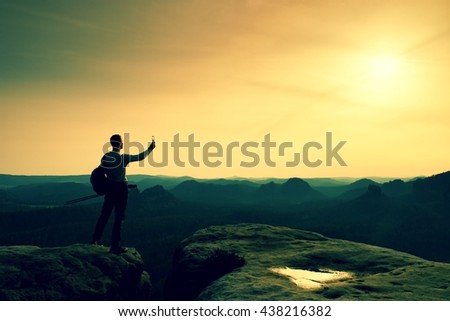 Tall tourist at dangerous cliff edge is taking selfie on peak above valley. Mountain peak and deep valley with sun in the frame
