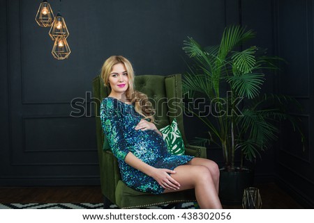 Young, extravagant, extraordinary pregnant girl, blonde woman sitting on a chair near palm tree in the flowerpot snake modern dress on a dark background with a green wall picture, embracing the belly