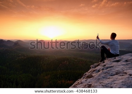 Tourist in grey t-shirt takes photos with smart phone on peak of rock. Dreamy hilly landscape below, spring orange pink misty sunrise in beautiful valley below rocky mountains. Hiking man taking photo