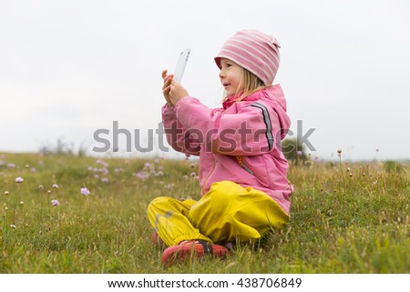 cute little girl in preschool age sitting on rural field with dry yellow grass and small flowers and playing with mobile phone, taking pictures or self portrait