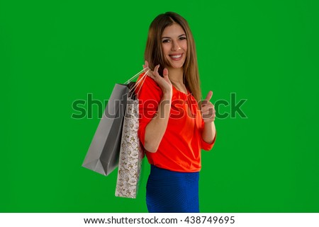 Cute young woman with shopping bags in hands smiling on camera and showing thumbs up isolated on green screen