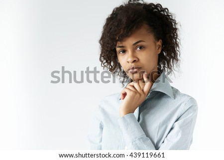 Young African female in striped shirt looking at the camera with serious pensive expression, touching her cheek with a finger, posing against white studio wall with copy space for your information