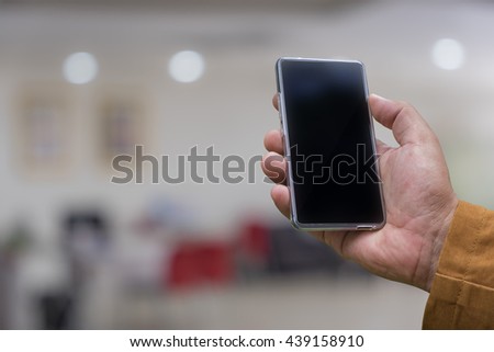 Man's hand shows mobile smartphone in vertical position, mockup template - abstract office blur background with bokeh.
