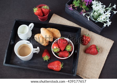Coffee, mini French pastries and strawberries on wooden tray over black table. White and purple flowers in a decorative wooden crate. Black background