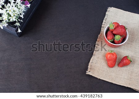 Fresh strawberries in mini metal bucket on hessian jute. White and purple flowers in a decorative wooden crate. Black background. 