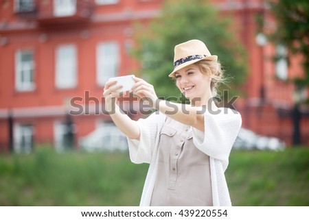 Happy beautiful woman traveler taking selfie with smartphone in front of sight location on the street. Young traveling female in straw hat making self-portrait with red building on the background