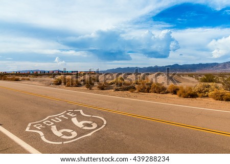 U.S. Route 66 highway, with sign on asphalt and a long train in the background, near amboy, california. Located in the mojave dessert Royalty-Free Stock Photo #439288234