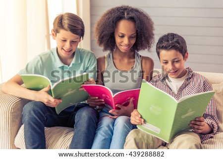 Three teenagers are reading and smiling while sitting on the couch at home. Attractive afro american girl is sitting between boys