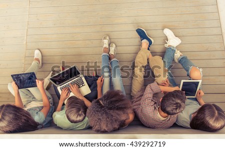 Top view of group of teenage boys and girls using gadgets while sitting in row on wooden floor Royalty-Free Stock Photo #439292719