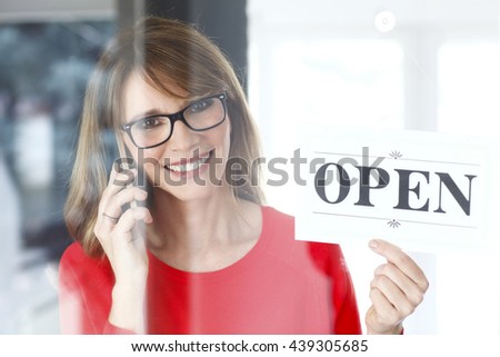 Portrait of a smiling middle aged woman making call while holding up a sign on opening day of her small business.