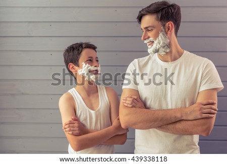 Handsome young father and his teenage son with shaving foam on their faces are looking each other and smiling, standing cross-armed against gray wall