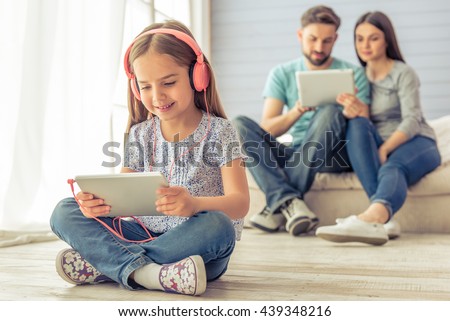 Cute little girl in headphones is using a tablet and smiling, in the background her parents are using a tablet too, sitting on sofa at home
