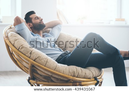 Total relaxation. Handsome young man keeping eyes closed and holding hands behind head while sitting in big comfortable chair at home   Royalty-Free Stock Photo #439431067