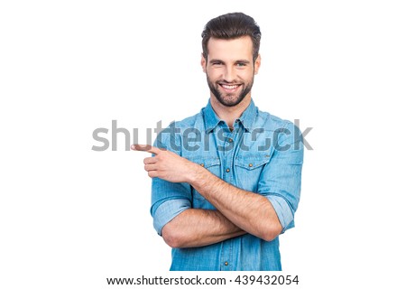 Look over there! Happy young handsome man in jeans shirt pointing away and smiling while standing against white background  Royalty-Free Stock Photo #439432054
